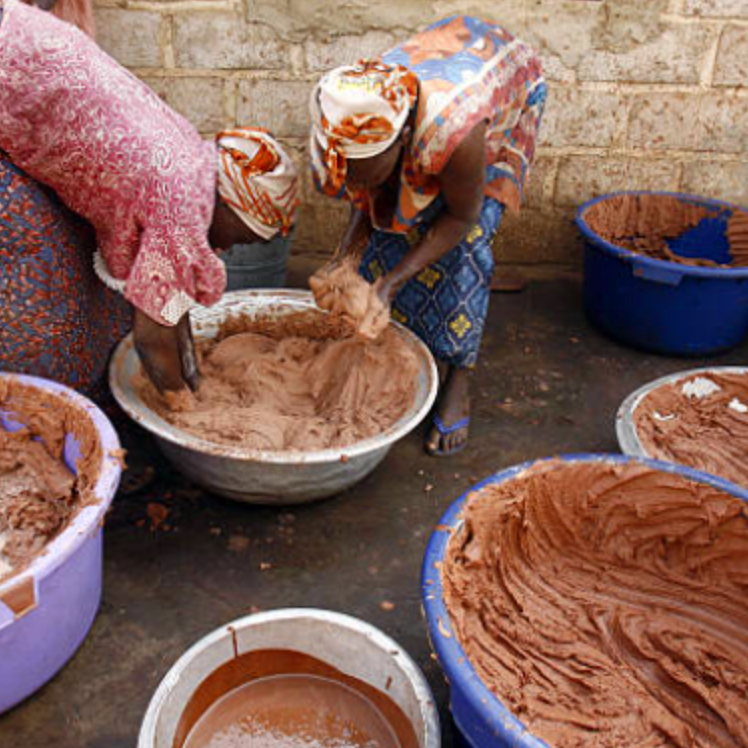 Woman mixing a brown shea butter substance in a bowl with other containers nearby part of the shea butter making process.